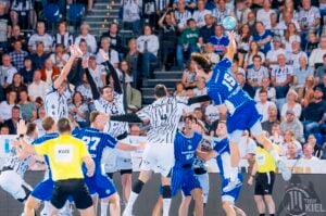 Ein Spieler in einem blauen Trikot springt hoch, um den Ball zu werfen, während Verteidiger in weiß gestreiften Trikots versuchen, ihn zu blockieren – und das alles bei einem Hallenhandballspiel vor einer großen Zuschauermenge.