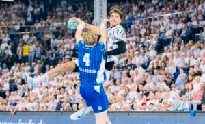 Ein Handballspieler in einem weißen Trikot springt hoch, um den Ball zu werfen, während er in der Luft von einem Verteidiger in einem blau gestreiften Trikot geblockt wird. Die Szene spielt sich vor ausverkauftem Haus in einer Halle ab.