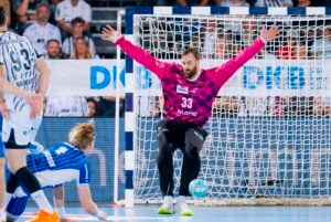 Ein Handballtorwart in einem rosa Trikot verteidigt das Tor mit weit ausgestreckten Armen und Beinen, während ein Spieler in Blau und Weiß den Ball schießt und die Zuschauer im Hintergrund zuschauen.