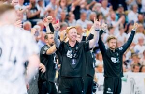 Eine Gruppe von Männern in schwarzen Sporttrikots jubelt mit erhobenen Armen, lächelt und feiert vor einer verschwommenen Menschenmenge bei einer Hallensportveranstaltung.