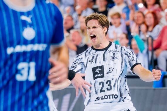 Ein Handballspieler im weißen Trikot mit der Nummer 23 jubelt begeistert auf dem Spielfeld. Im Vordergrund ist ein weiterer Spieler im blauen Trikot mit der Nummer 31 zu sehen. Im Hintergrund ist das Publikum zu sehen, das jubelt.