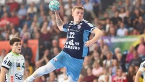 Ein Handballspieler in einem blauen Hamburger Trikot springt mit dem Ball in der Hand in die Luft und will gerade schießen. Ein anderer Spieler in einem weißen Trikot schaut zu. Die Zuschauer im Hintergrund sind verschwommen.