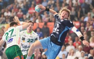Ein Handballspieler in einem marineblauen und hellblauen Trikot springt hoch, um den Ball zu werfen, während zwei Spieler in Weiß und Grün versuchen, ihn während eines Spiels zu blocken; im Hintergrund ist eine verschwommene Zuschauermenge zu sehen.