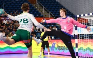 Ein Handballspieler in einem grün-weißen Trikot springt hoch, um einen Wurf abzugeben, während sich ein Torwart in Rosa streckt, um den Wurf abzuwehren. Die Szene spielt in einer Sporthalle, im Hintergrund sind Zuschauer zu sehen.