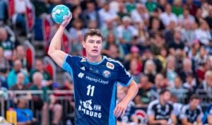 Ein männlicher Handballspieler in einem blauen Trikot mit der Nummer 11 macht sich während eines Spiels bereit, den Ball zu werfen. Im Hintergrund sind die verschwommenen Umrisse der Zuschauer und anderer Spieler zu erkennen.