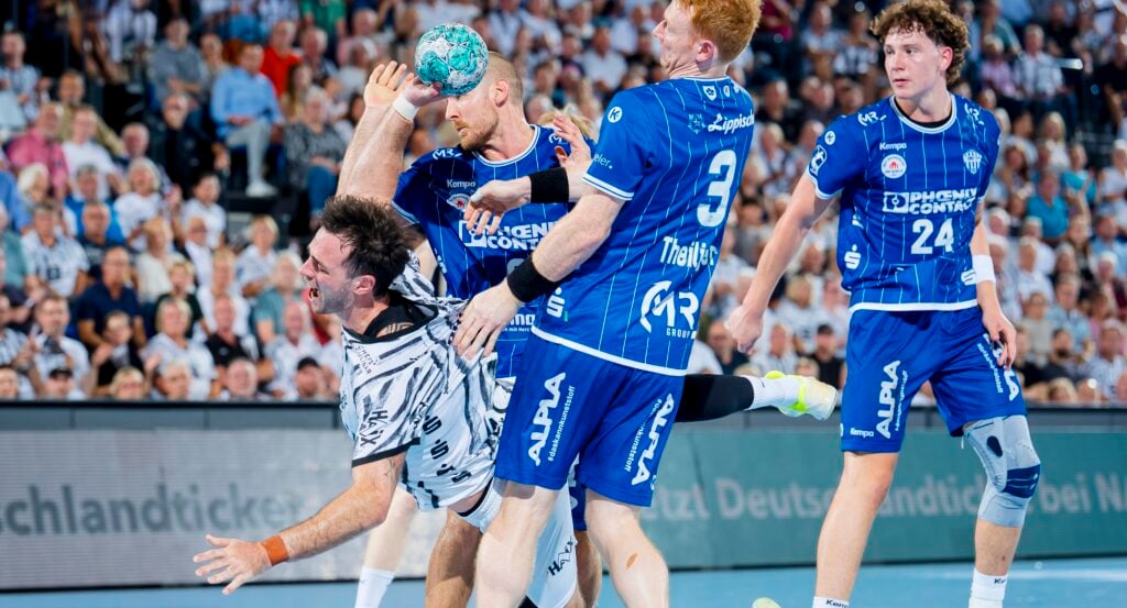 Ein Handballspieler in einem weißen Trikot wird während eines Spiels von Gegnern in blauen Trikots gefoult, während die Zuschauer von den Tribünen aus zusehen.