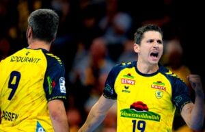 Zwei männliche Sportler in gelben Trikots – der eine mit dem Rücken zur Kamera, der andere mit geballter Faust und laut rufend – stehen auf einem Hallensportplatz; ihre Trikots sind mit Sponsorenlogos und Rückennummern versehen.