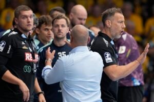 Ein Handballtrainer in einem hellblauen Hemd deutet während eines Spiels auf einen Spieler in einem schwarzen Mannschaftstrikot.