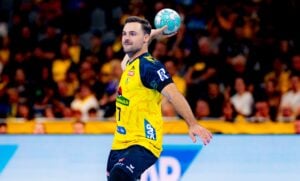 Ein männlicher Handballspieler in einem gelb-schwarzen Trikot macht sich während eines Spiels bereit, den Ball zu werfen; im Hintergrund ist eine Zuschauermenge zu sehen.