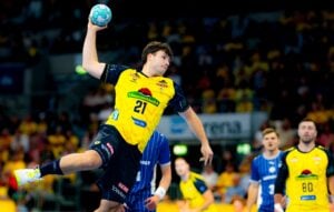 Ein männlicher Handballspieler in einem gelben Trikot springt in der Luft hoch, um den Ball zu werfen, während Spieler in blauen Trikots bei einem Spiel in einer Sporthalle zuschauen.