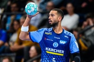 Ein Handballspieler in einem blauen Trikot hält einen Handball in der rechten Hand hoch.