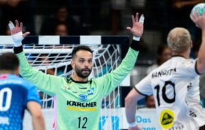 Ein Handballtorwart in einem hellgrünen Trikot steht mit erhobenen Armen vor dem Tor und blickt auf einen Gegner, der gerade den Ball werfen will.