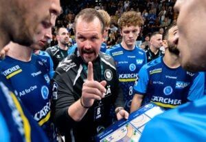 Ein Handballtrainer in einer schwarzen Jacke gestikuliert und spricht mit seiner Mannschaft, die sich um ihn versammelt hat und ihm aufmerksam zuhört, während er eine taktische Tafel in der Hand hält.