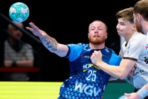 Ein Handballspieler im blauen Trikot macht sich bereit, den Ball zu werfen, während er von einem Gegner im weißen Trikot verteidigt wird.