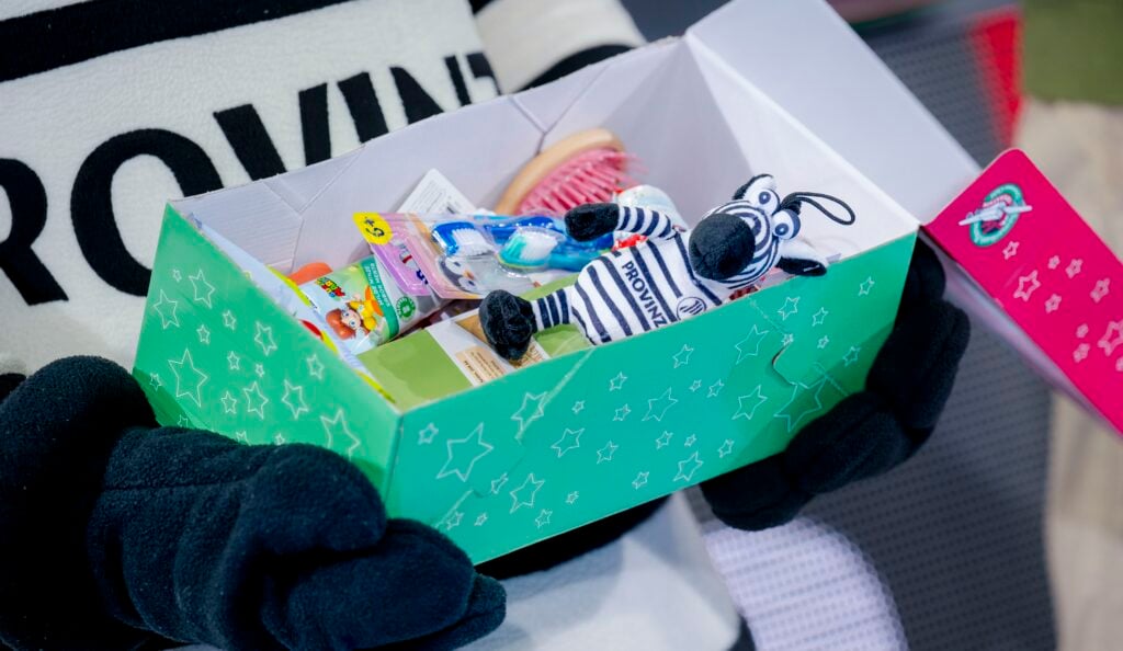 HANDBALL 2025/26: THW Kiel - Bergischer HC Eine Person mit schwarzen Handschuhen hält eine grüne Geschenkbox in der Hand, die mit Toilettenartikeln für Kinder und einem kleinen Zebra-Plüschtier gefüllt ist.