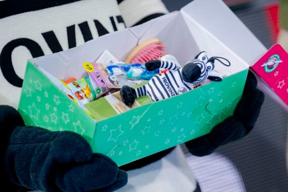 Eine Person mit schwarzen Handschuhen hält eine grüne Geschenkbox in der Hand, die mit Toilettenartikeln für Kinder und einem kleinen Zebra-Plüschtier gefüllt ist.