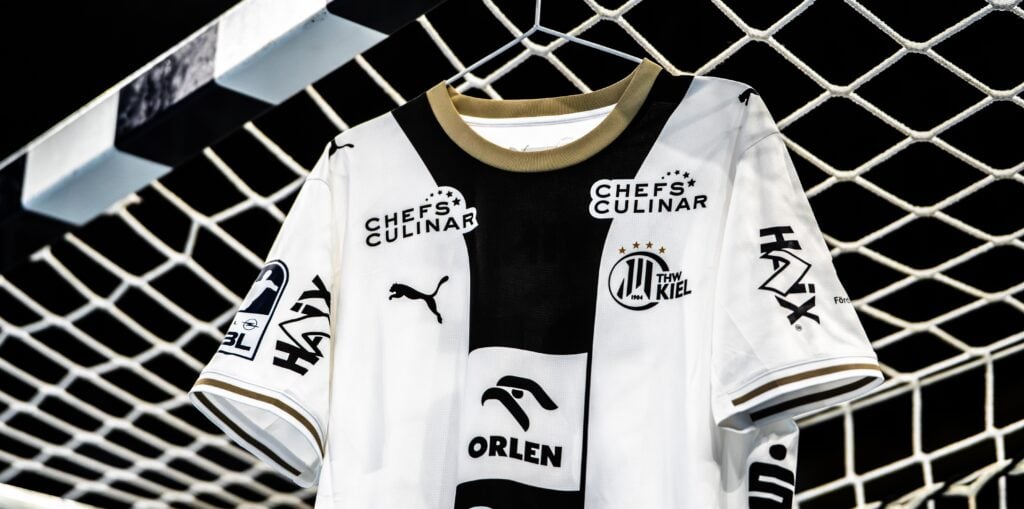 Weißes THW Kiel-Handballtrikot mit schwarzen Akzenten und Sponsorenlogos, das vor einem Tornetz hängt.