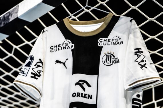 Weißes THW Kiel-Handballtrikot mit schwarzen Akzenten und Sponsorenlogos, das vor einem Tornetz hängt.