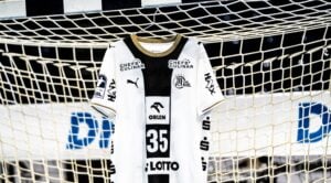 Ein weißes Handballtrikot des THW Kiel mit schwarzen und goldenen Akzenten, das im Tornetz hängt.