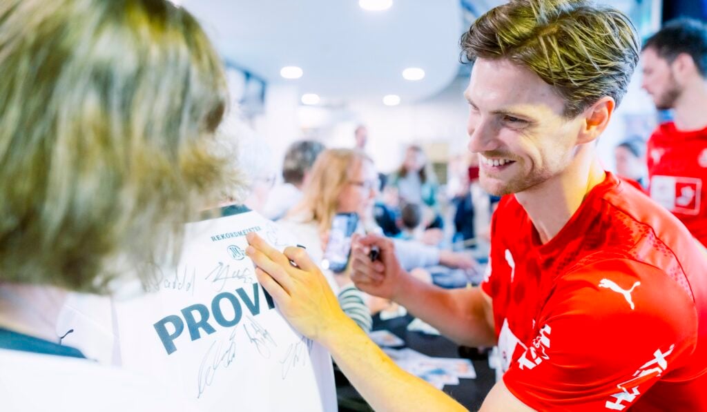 HANDBALL 2025/26: THW Kiel - TBV Lemgo Lippe Ein lächelnder Sportler in einem roten T-Shirt signiert bei einer Hallenveranstaltung ein weißes Trikot für einen Fan. Im Hintergrund sind weitere Personen und Sportler zu sehen.