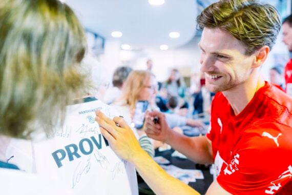 Ein lächelnder Sportler in einem roten T-Shirt signiert bei einer Hallenveranstaltung ein weißes Trikot für einen Fan. Im Hintergrund sind weitere Personen und Sportler zu sehen.