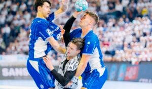 Drei Sportler kämpfen während eines Handballspiels um den Ball; zwei Spieler in Blau blockieren einen Spieler in Weiß und Schwarz, im Hintergrund ist eine verschwommene Zuschauermenge zu sehen.