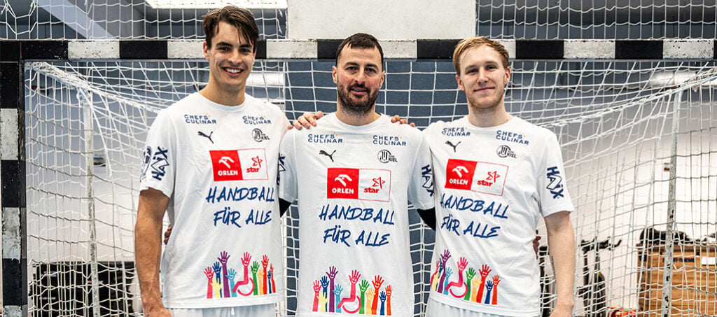 Header_Inklusions-Shirt Drei Handballspieler stehen vor einem Tor und tragen weiße Trikots mit der Aufschrift HANDBALL FÜR ALLE und bunten Hand-Illustrationen, um beim Heimspiel des THW Kiel gegen Stuttgart für Inklusion zu werben.