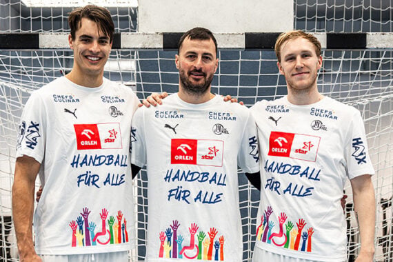 Drei Handballspieler stehen vor einem Tor und tragen weiße Trikots mit der Aufschrift HANDBALL FÜR ALLE und bunten Hand-Illustrationen, um beim Heimspiel des THW Kiel gegen Stuttgart für Inklusion zu werben.