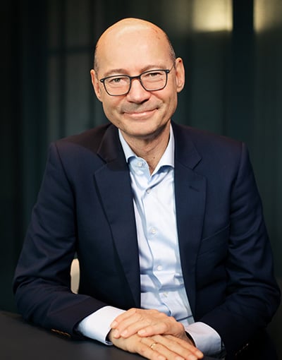 Wirtschaftsausschuss_Bilder_wernerWerner Ein Mann mittleren Alters mit Brille, der einen marineblauen Blazer und ein hellblaues Hemd trägt, sitzt an einem Tisch und lächelt als Mitglied der Geschäftsführung des THW Kiel.
