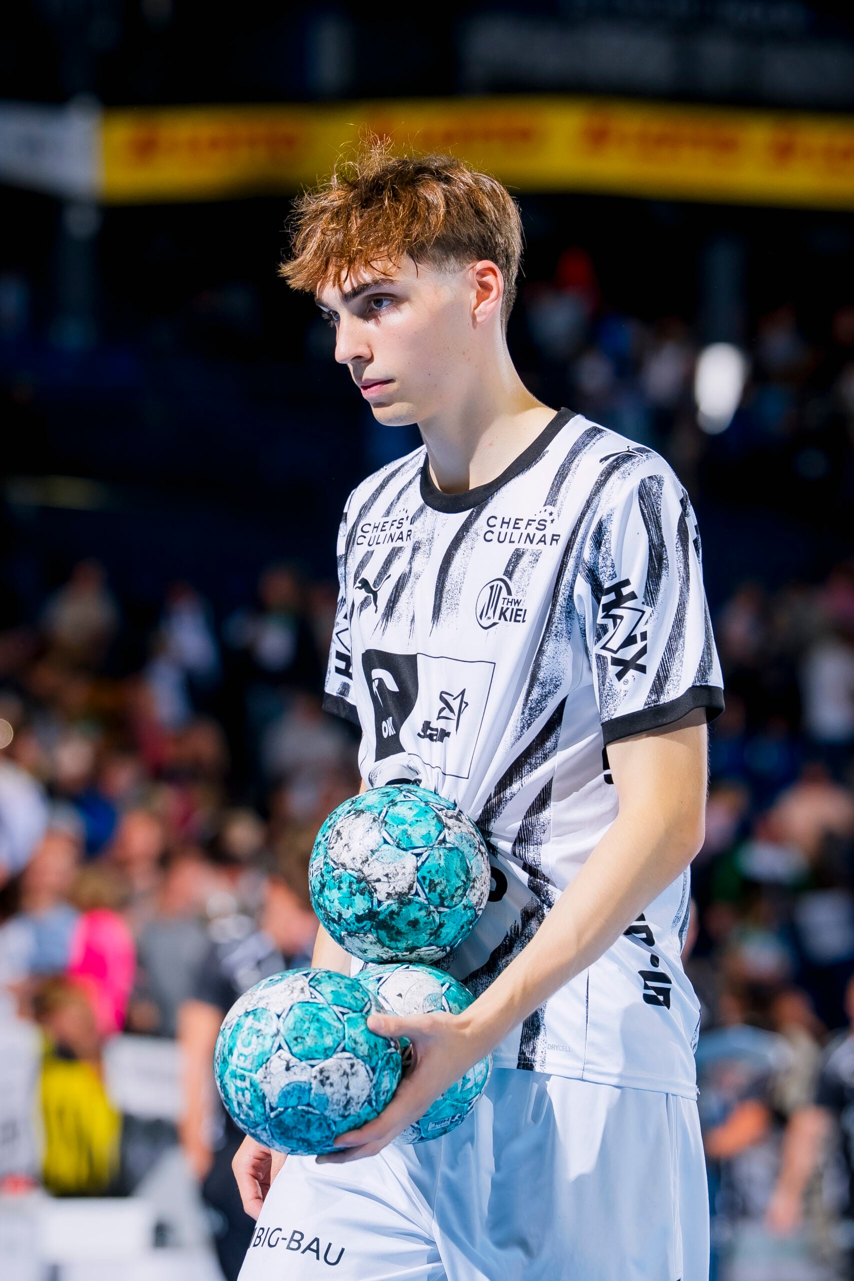Eric Johansson, ein junger Handballspieler des THW Kiel, hält zwei blau-weiß gemusterte Handbälle in den Armen.