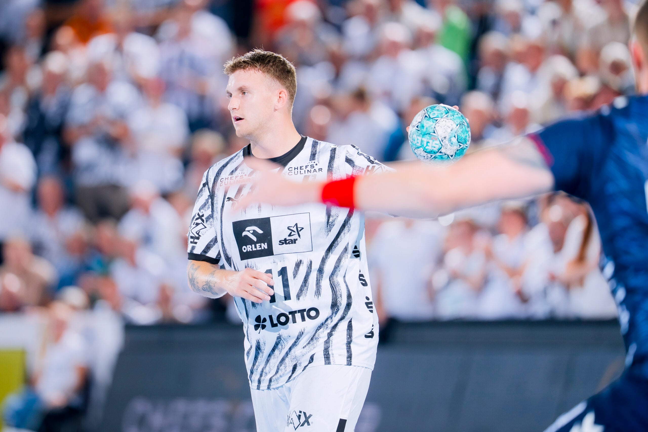 Eric Johansson, der ein weißes, gemustertes Trikot des THW Kiel trägt, macht sich bereit, während eines Handballspiels in einer vollbesetzten Halle einen blau-weißen Ball zu werfen.