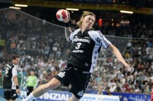 Ein Handballspieler des THW Kiel in schwarz-weißem Trikot springt in die Luft und macht sich bereit, den Ball zu werfen – während der neuen Handball-Bundesliga-Saison 2026/2027, während der neue Trainer Börge Lund am Spielfeldrand inmitten jubelnder Zuschauer zusieht.